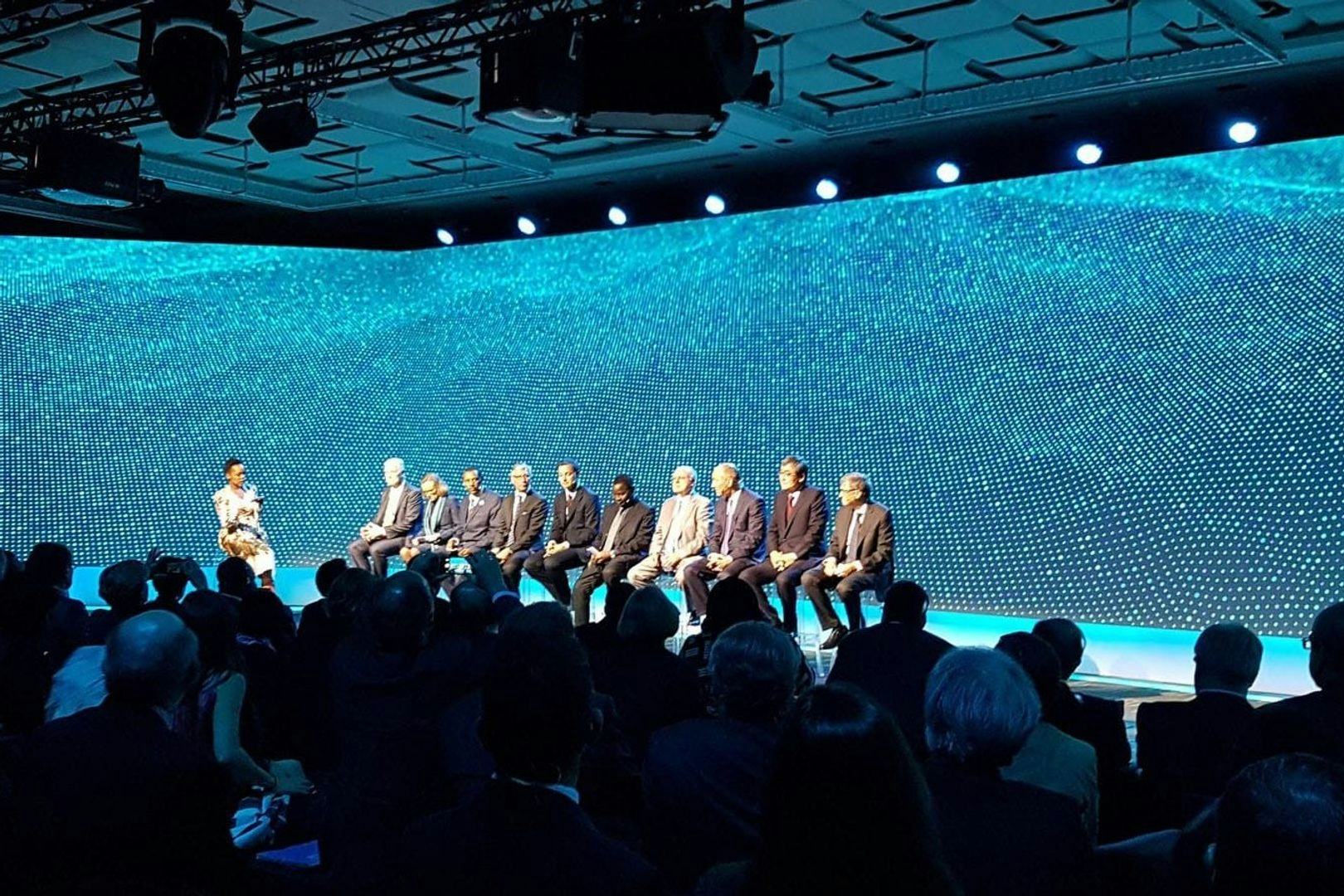 A photo taken during the Cascade presentation. Ten speakers sit on stage in front of three floor to ceiling blue screens that display a mesh moving like the surface of water. In the foreground, an audience sit in the dark watching the stage.